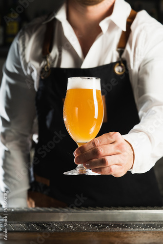 Beer. A professional bartender in a white shirt and black apron holds a glass of beer, the trend drinks concept. Vertical photo 