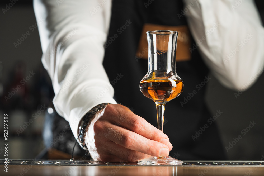 Strong alcohol. A professional bartender in a white shirt and black apron holds a tasting glass, the trend drinks concept.