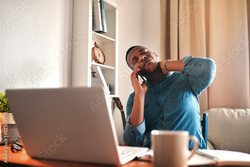 Tired business man with neck pain on a laptop and phone call, looking stressed and stretching bad, strained muscle or sore back. Stressed, multitasking guy having a difficult time working from home photo