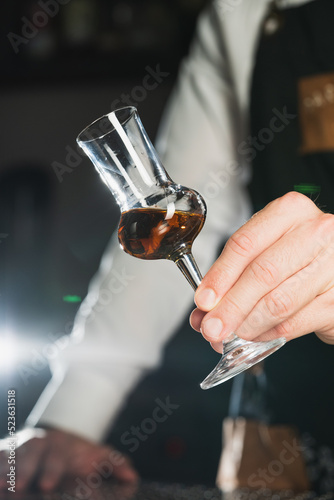 Strong alcohol. A professional bartender in a white shirt and black apron holds a tasting glass, the trend drinks concept. Vertical photo 