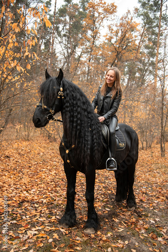 young beautiful smiling woman in black dress riding black Friesian horse in autumn forest with yellow leaves  © Tetatet