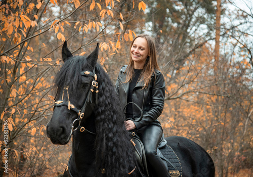 young beautiful smiling woman in black gothic dress riding black Friesian horse  in autumn forest with yellow leaves © Tetatet