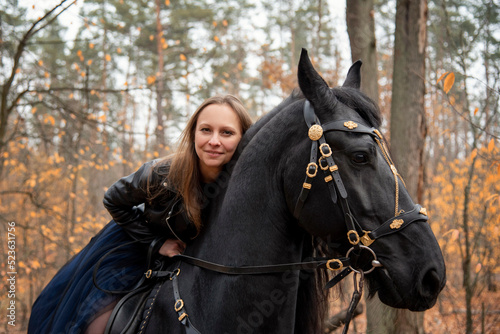 young beautiful smiling woman in long blue skirt riding black Friesian horse in autumn forest with yellow leaves