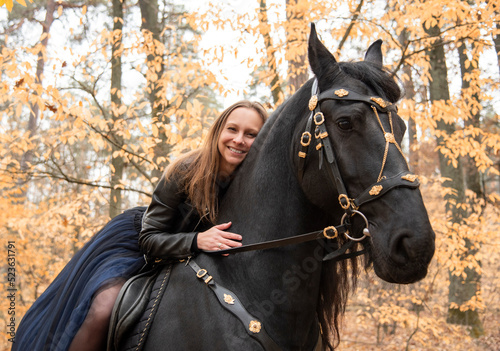 young beautiful smiling woman in long blue skirt riding black Friesian horse in autumn forest with yellow leaves