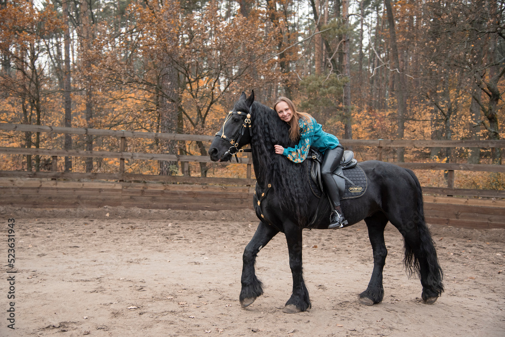 young beautiful smiling woman in ukrainian green embroidery riding black Friesian horse on a farm in autumn young beautiful smiling woman in ukrainian green embroidery riding black Friesian horse on a farm in autumn