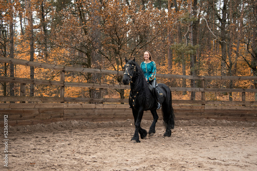 young beautiful smiling woman in ukrainian green embroidery riding black Friesian horse on a farm in autumn © Tetatet