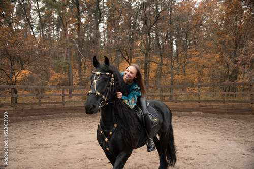 young beautiful smiling woman in ukrainian green embroidery riding black Friesian horse on a farm in autumn