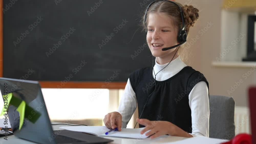 Smiling cute smart girl in headphones waving greeting watching online chat on laptop talking. Positive Caucasian intelligent schoolgirl studying remotely indoors. Modern technologies and education Smiling cute smart girl in headphones waving greeting watching online chat on laptop talking. Positive Caucasian intelligent schoolgirl studying remotely indoors. Modern technologies and education