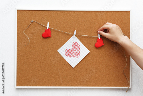 A woman's hand attaches a clothespin with a red heart drawn on a piece of paper to a cork board, space for text.