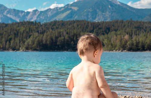 naked adorable baby boy sitting with back on scenic beautiful mountains lake landscape background playing in water.rocks pebbles beach turquoise blue clean water.sunny summer day light.kid having fun