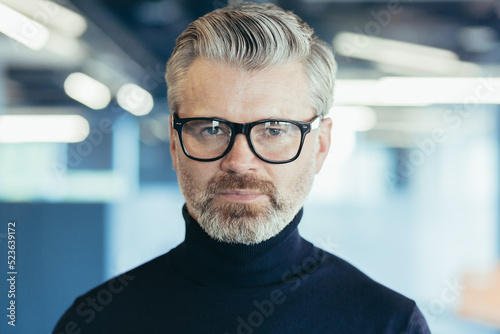 Close-up photo portrait of successful serious and mature businessman, investor in glasses looking thoughtfully at camera, man at work inside office