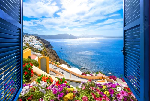 Fototapeta View from an open window with shutters and flowerbox, over the gardens and terraces of Santorini Greece and the blue Aegean Sea.