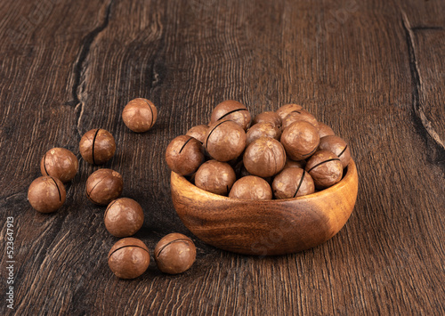 Unpeeled macadamia nuts in a bowl and on a wooden background.