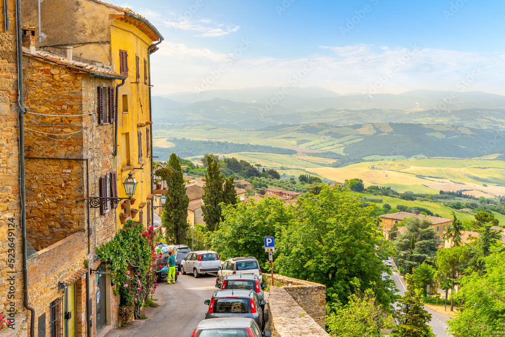 Cars park along the narrow street below stone homes overlooking the hills of Tuscany, on the outer wall of the walled medieval village of Volterra, Italy.