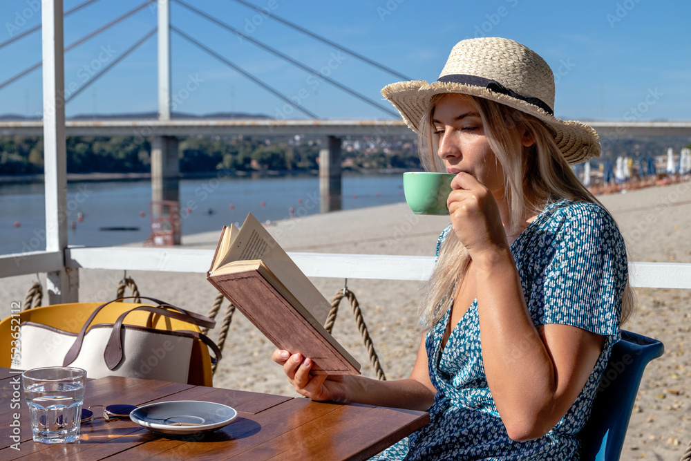 A beautiful young woman with a straw hat enjoys a lovely summer afternoon, a good book, and a cup of coffee in a cafe by the river.