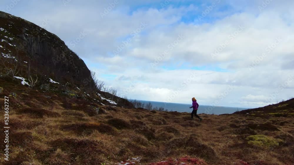 Panning Shot Of Female Tourist In Warm Clothing Running On Mountain By Sea During Vacation - Tromso, Norway Panning Shot Of Female Tourist In Warm Clothing Running On Mountain By Sea During Vacation - Tromso, Norway
