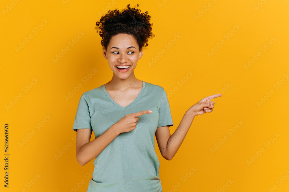 Excited curly haired young african american woman in a basic t-shirt, with positive emotional facial expression, stand on isolated orange background, looks and points fingers to the side, smiling Excited curly haired young african american woman in a basic t-shirt, with positive emotional facial expression, stand on isolated orange background, looks and points fingers to the side, smiling