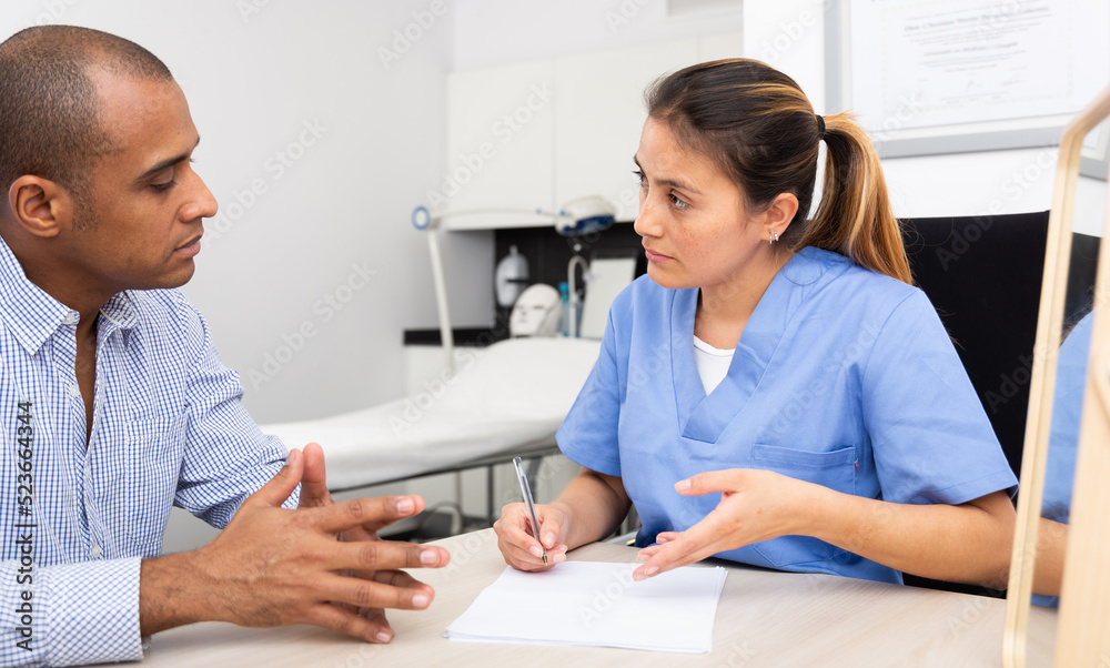 Fototapeta premium Young polite Latina cosmetologist doctor working with male client in clinic, filling out medical form before procedure ..