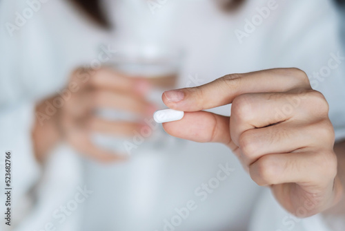 Adult woman holding pill and glass of water, female taking medicine on bed at home. Migraine, painkiller, headache, influenza, illness, sickness and healthcare concept