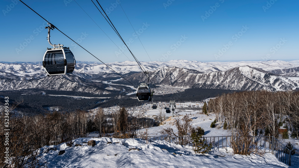 Obraz premium Cable car cabins on ropes over a snow-covered valley. A picturesque mountain range against the background of the blue sky in the distance. Altai. Manzherok Ski Resort