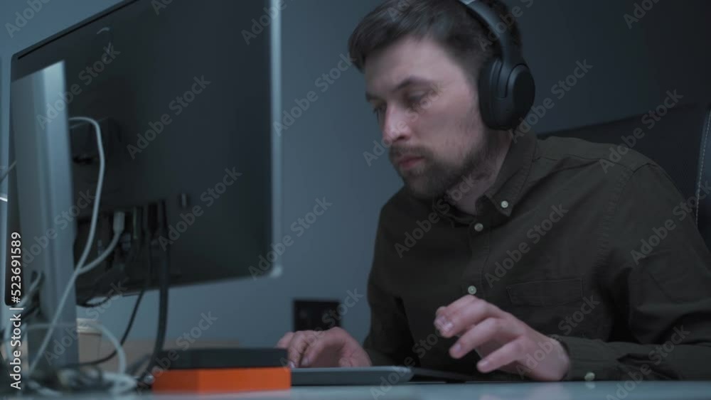 Male freelancer in shirt, working at home at computer in night. Wearing headphones, typing on keyboard and looking at monitor. IT engineer in headphones working on computer in office at late evening.