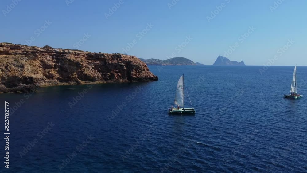 Catamaran with white sails in a beautiful bayTranquil aerial view flight fly backwards drone footageof cliff edge beach Sa Figuera borda Ibiza summer 2022.