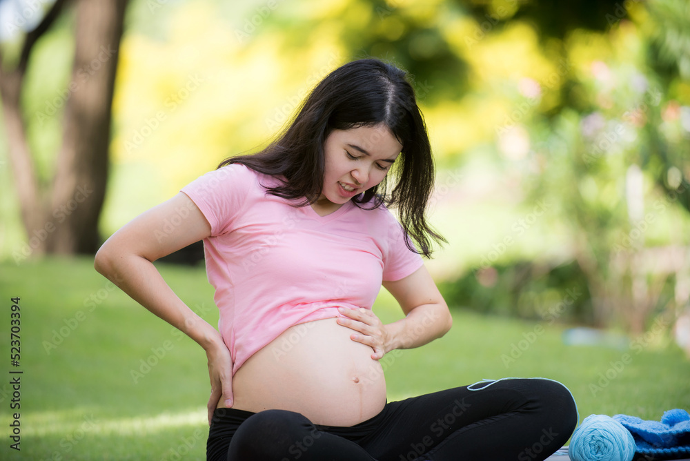 Asian pregnant woman suffering from back pain A beautiful pregnant woman holds her belly and puts one hand behind her back while sitting in the park. Asian pregnant woman suffering from back pain A beautiful pregnant woman holds her belly and puts one hand behind her back while sitting in the park.