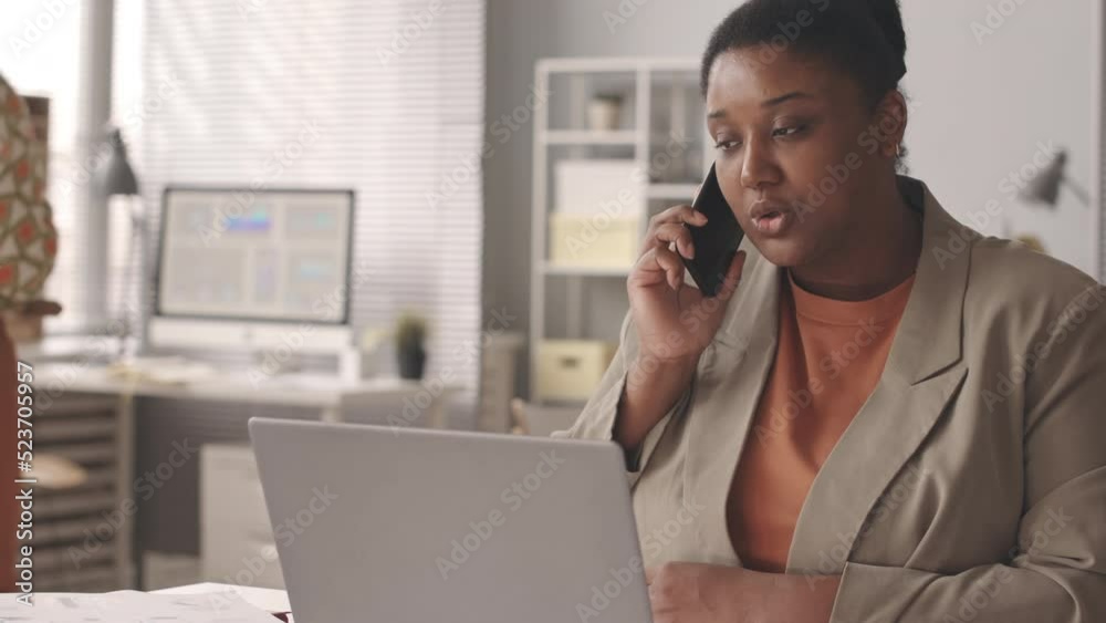 Waist up slowmo of young Black woman talking on mobile phone as sitting in front of laptop at desk in office, working there with female colleague