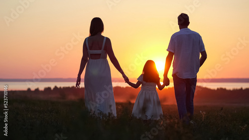 Happy parents silhouettes with excited daughter walk on evening meadow. Family looks together at bright sunset walking on lawn against river