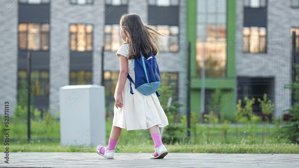 Preschooler girl with blue backpack walks past big school building. Schoolkid goes to school hopping in evening. Girl wearing white dress enjoys going to school