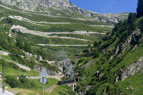 Look up to serpentine mountain road of Swiss Grimsel Pass with smoke of Furka steam train on a sunny summer day. Photo taken July 3rd, 2022, Oberwald, Switzerland.