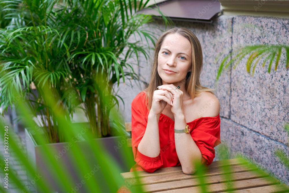 A confident young woman sits at a table in a street cafe in a red dress, waiting for her order. Caucasian woman came to breakfast A confident young woman sits at a table in a street cafe in a red dress, waiting for her order. Caucasian woman came to breakfast