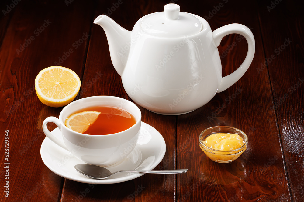 Tea with lemon and honey in a porcelain cup next to a white teapot on a wooden table background. Angle view. Selective focus. Tea with lemon and honey in a porcelain cup next to a white teapot on a wooden table background. Angle view. Selective focus.