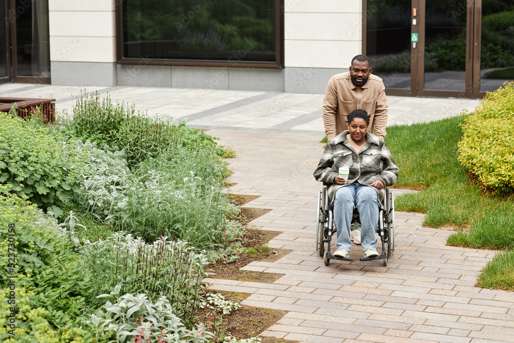 Full length portrait of modern black couple with smiling young woman in wheelchair enjoying walk in city garden, copy space Full length portrait of modern black couple with smiling young woman in wheelchair enjoying walk in city garden, copy space