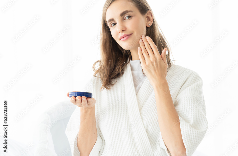 Woman caring of her beautiful skin on the face applying moisturizer on her face. Smiling girl holding little jar of skin cream and applying lotion after shower Woman caring of her beautiful skin on the face applying moisturizer on her face. Smiling girl holding little jar of skin cream and applying lotion after shower