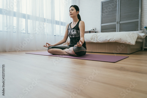low angle shot asian woman in sportswear is sitting in meditative pose with mudra hand signs on yoga mat ready to practicing lotus pose in the bedroom at home.