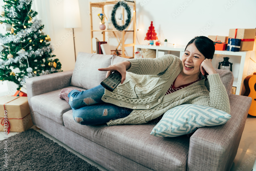 Obraz premium asian female couch potato is pointing at screen while watching funny tv shows. woman lying on sofa on her side is having a hearty laugh. indoor recreation ideas for Christmas holiday. real moments