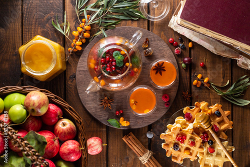 Autumn tea with berries and honey. Sea buckthorn and raspberries in a glass teapot, cups with poured tea. Apples and waffles with honey.