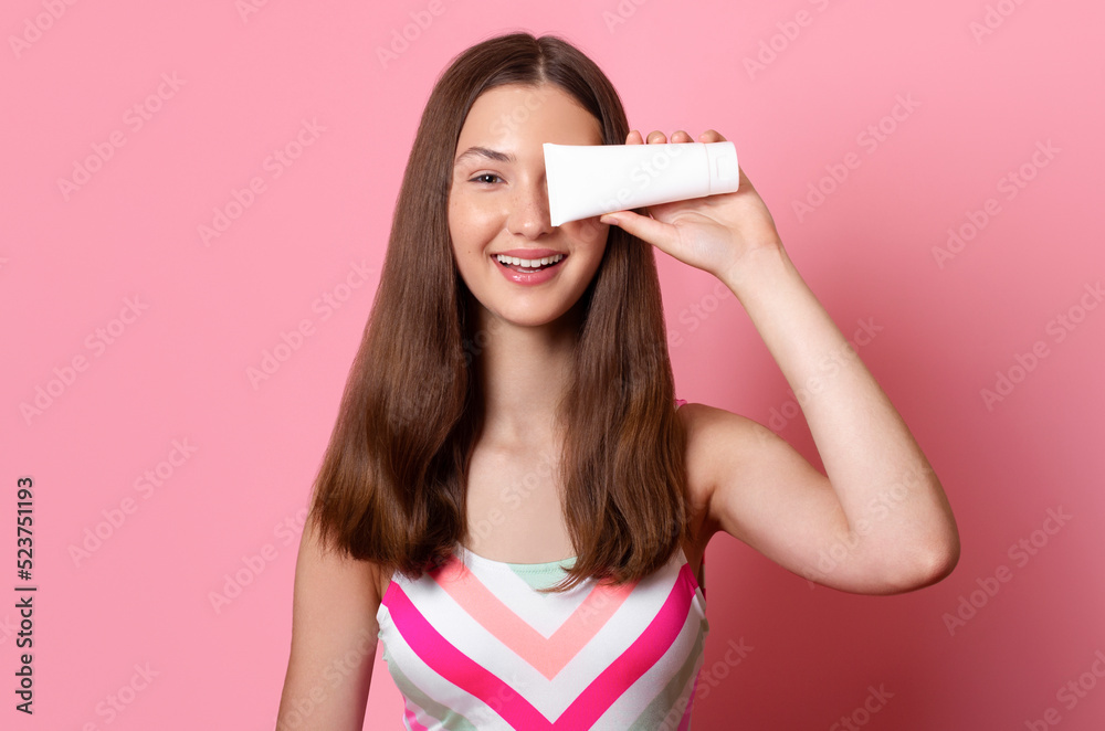 Cheerful smiling tanned teen girl hold in hand white blank cream tube mock up sunscreen, wearing colorful one piece swimsuit isolated on pink background Cheerful smiling tanned teen girl hold in hand white blank cream tube mock up sunscreen, wearing colorful one piece swimsuit isolated on pink background