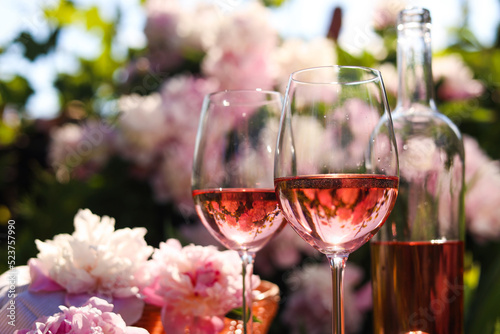 Fototapeta Naklejka Na Ścianę i Meble -  Bottle and glasses of rose wine near beautiful peonies in garden, closeup. Space for text