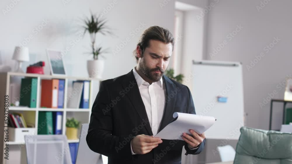 Thoughtful man in suit carefully reading important documents on clipboard in modern office room. Caucasian male director analyzing expenses on new business idea. Thoughtful man in suit carefully reading important documents on clipboard in modern office room. Caucasian male director analyzing expenses on new business idea.