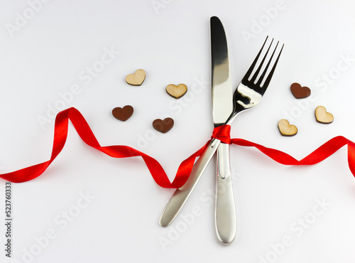 Romantic dinner concept. Top view of silver fork and knife decorated red ribbon with bow and wooden hearts on white background