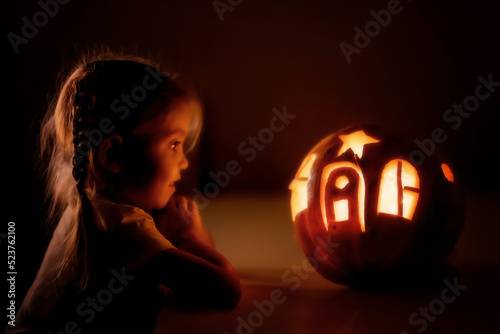 Happy excited little girl looking inside of glowing jack o lantern, carved pumpkin, while standing in dark house room decorated for all hallows eve. Halloween day, selective focus photo