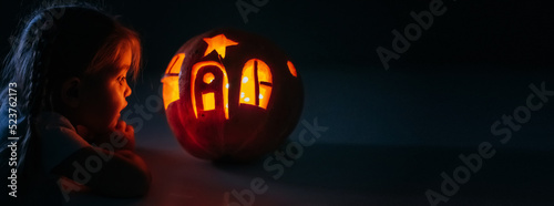 Quadro su tela Happy excited little girl looking inside of glowing jack o lantern, carved pumpkin, while standing in dark house room decorated for all hallows eve