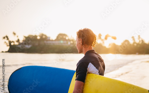 Young teenager boy with colorful surfboard go to the sea for surfing. He enjoying a beautiful sunset light on Dewata beach on Sri Lanka. Active sport vacation and exotic countries traveling concept.