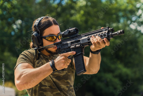 White muscular man in camouflage t-shirt and safety gear firing submachine gun on training ground. Outdoor horizontal shot. High quality photo