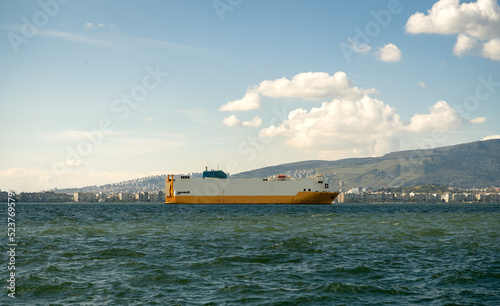 White and yellow ship cruising on the sea. City landscape behind it with cloudy sky.