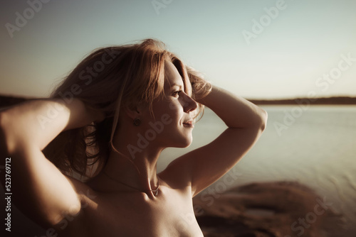Young woman of European appearance with tanned skin is posing nude looking into distance on the sea beach during the sunset. Beautiful girl portrait. Sexy, playful look. Liberated human no complexes.