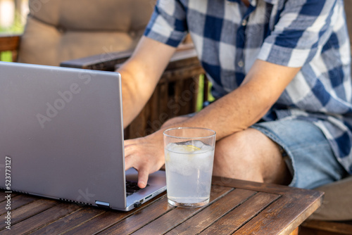 Side view, male hands type on a laptop on a wooden table, next to a glass with a refreshing drink, in a relaxed atmosphere.