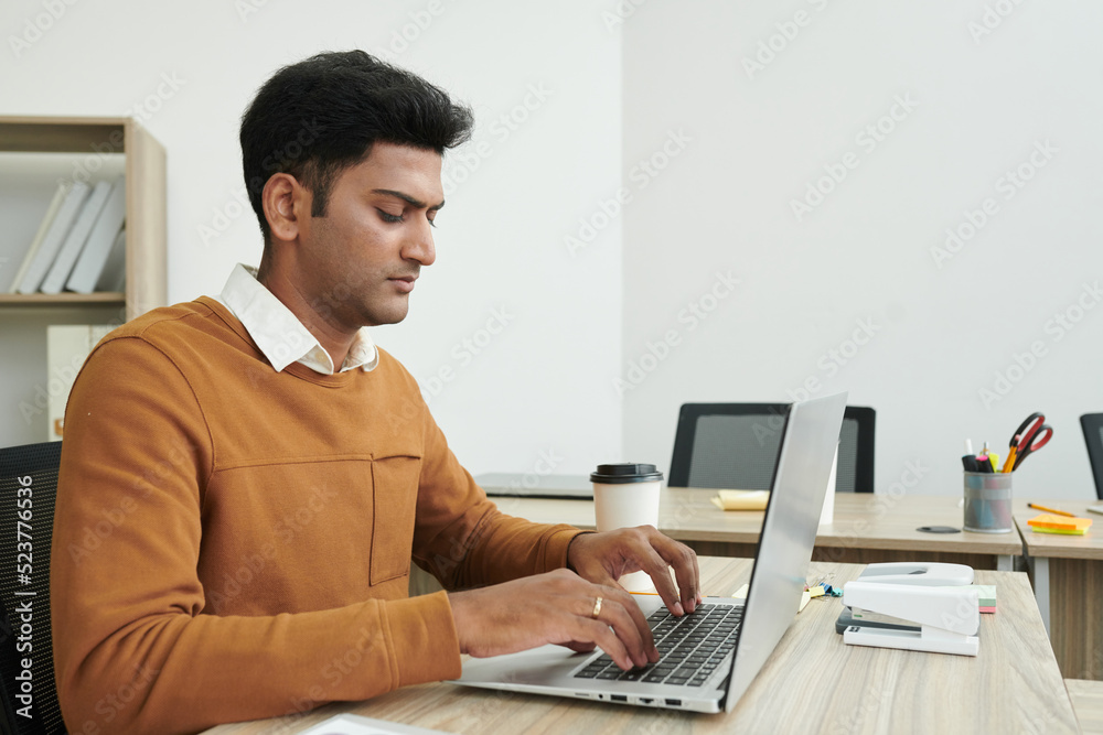 Serious young Indian businessman working on laptop at office desk Serious young Indian businessman working on laptop at office desk